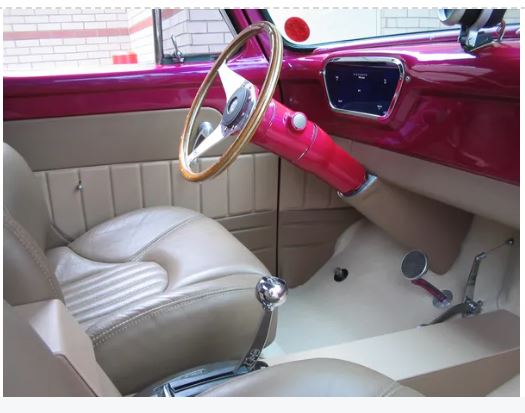 Interior view of a vehicle featuring a custom steering column cover in a pinkish hue, with a tan steering wheel and seat.