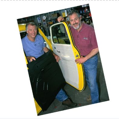 Two men holding a car door and a black panel in a workshop setting